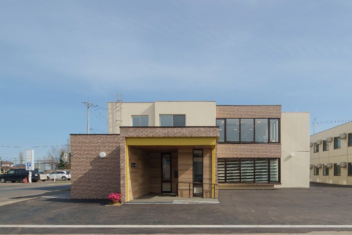 青い空の下に建っている2階建ての建物はグレー系の外壁のシンプルなデザインで、建物の周りにはプランターがある。