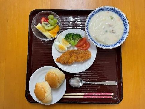 ロールパンにカットフルーツの盛り合わせ、野菜や卵が添えられたフライ、クリームシチューが並んでいる。