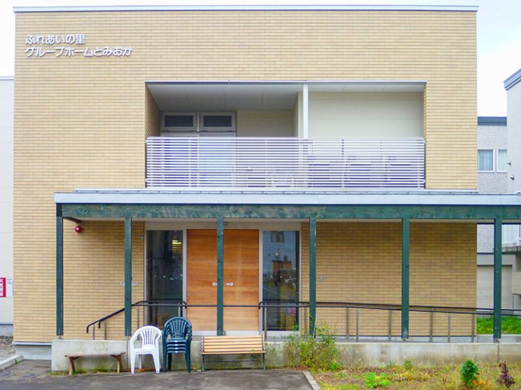 2階建ての建物はベージュ系の外観で、玄関には屋根と手すり付きのスロープがあり、いすが置かれている。
