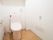 トイレには手洗い管の付いた便器が設置されており、床にはトイレブラシや芳香剤が置かれている。右の壁には呼び出しボタンが備えられている。