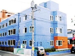 ブルーの大きな建物は3階建てで、駐車スペースもある。建物の近くにはグリーンのネットがかけられたごみステーションもある。