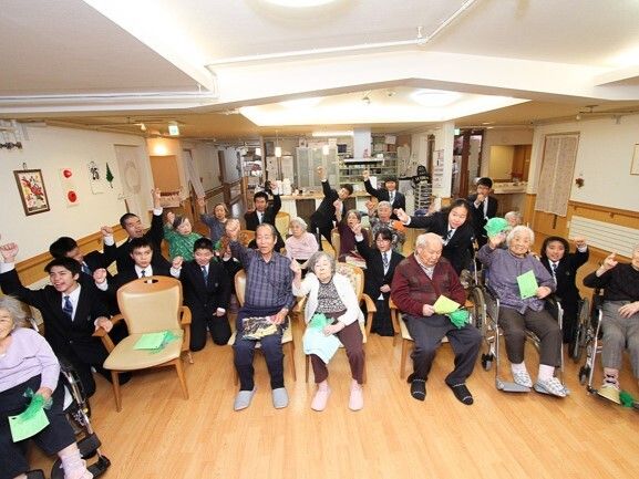 フローリングの共有スペースに集まり、高齢者たちとスーツ姿の若者たちが片手をグーにしてあげている。