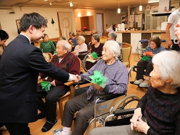 車いすに座った高齢女性がスーツ姿の若い男性と笑顔で握手をしている様子。