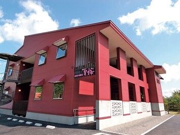 緑に囲まれた場所にある赤い外壁の２階建ての建物は、玄関から緩やかなスロープが付いている。