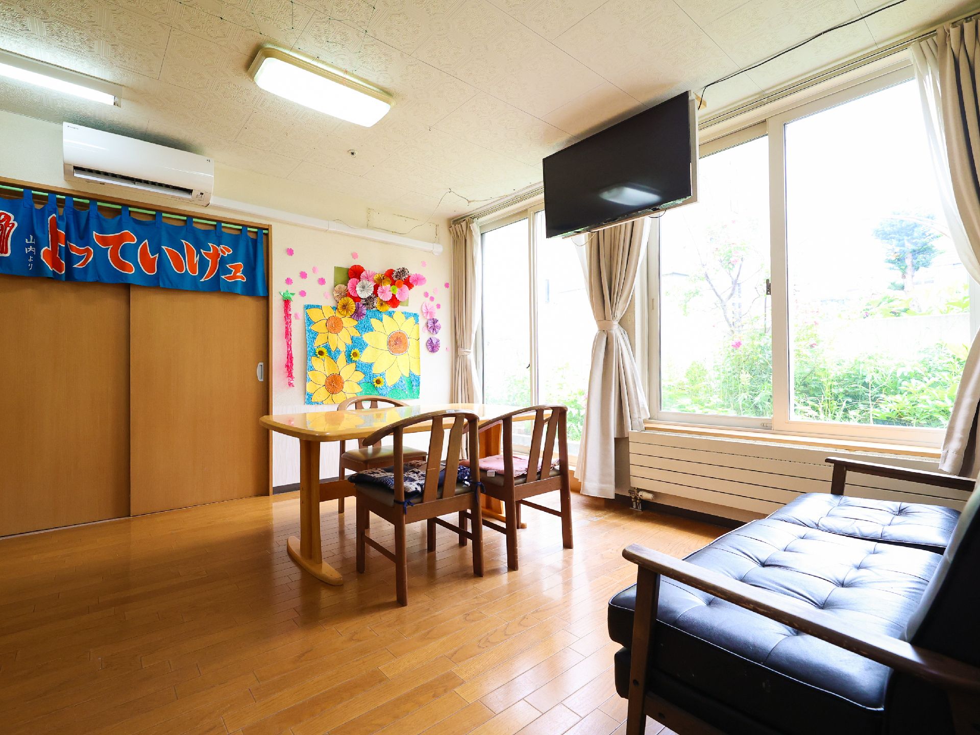壁一面が大きな窓になっているテレビが天井から下げられ机と椅子とソファーがある部屋
