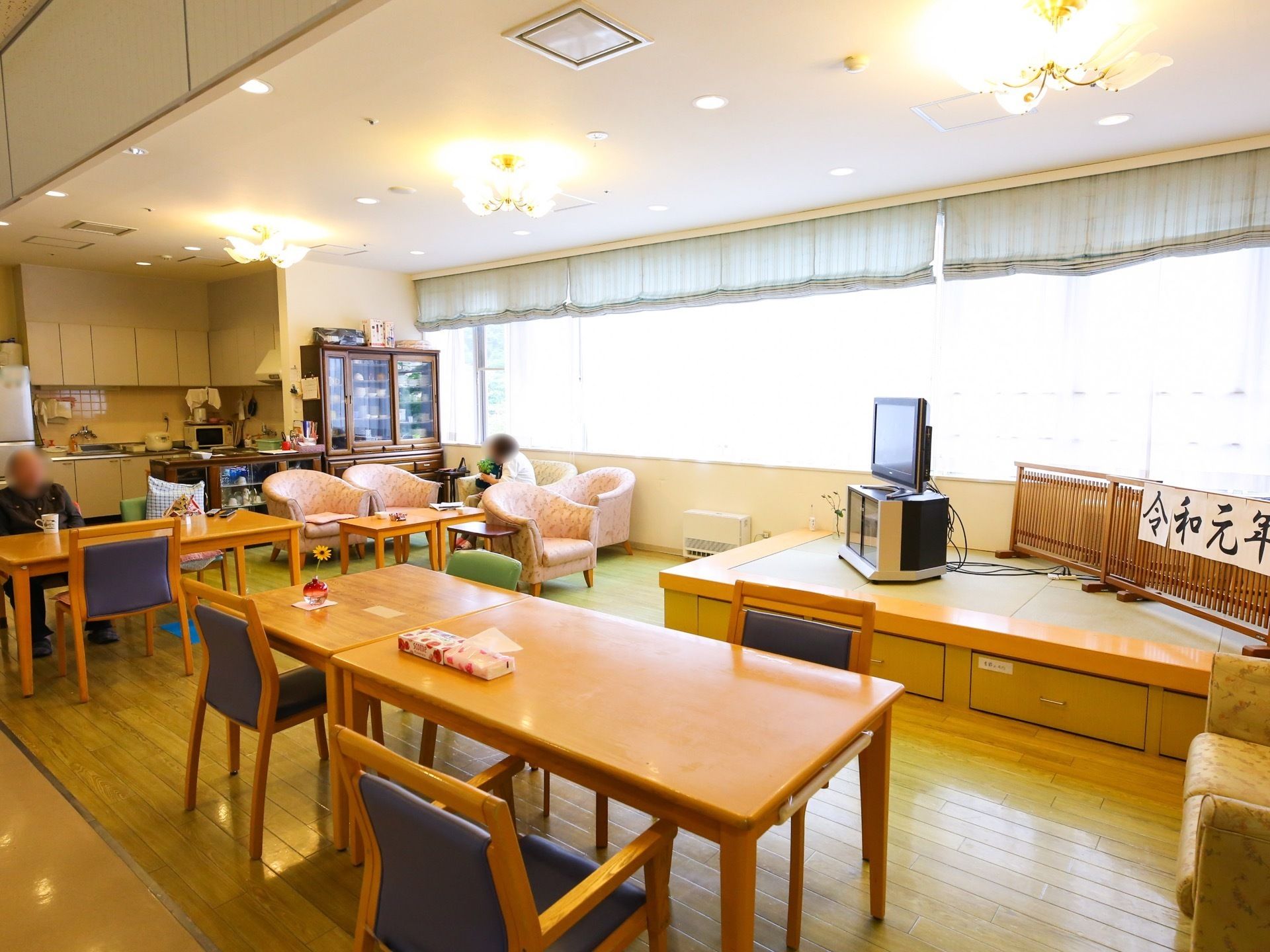 窓が大きく設置されており、ロールカーテンが付いている。壁際にはキッチンがあり、付近には冷蔵庫や食器棚がある。