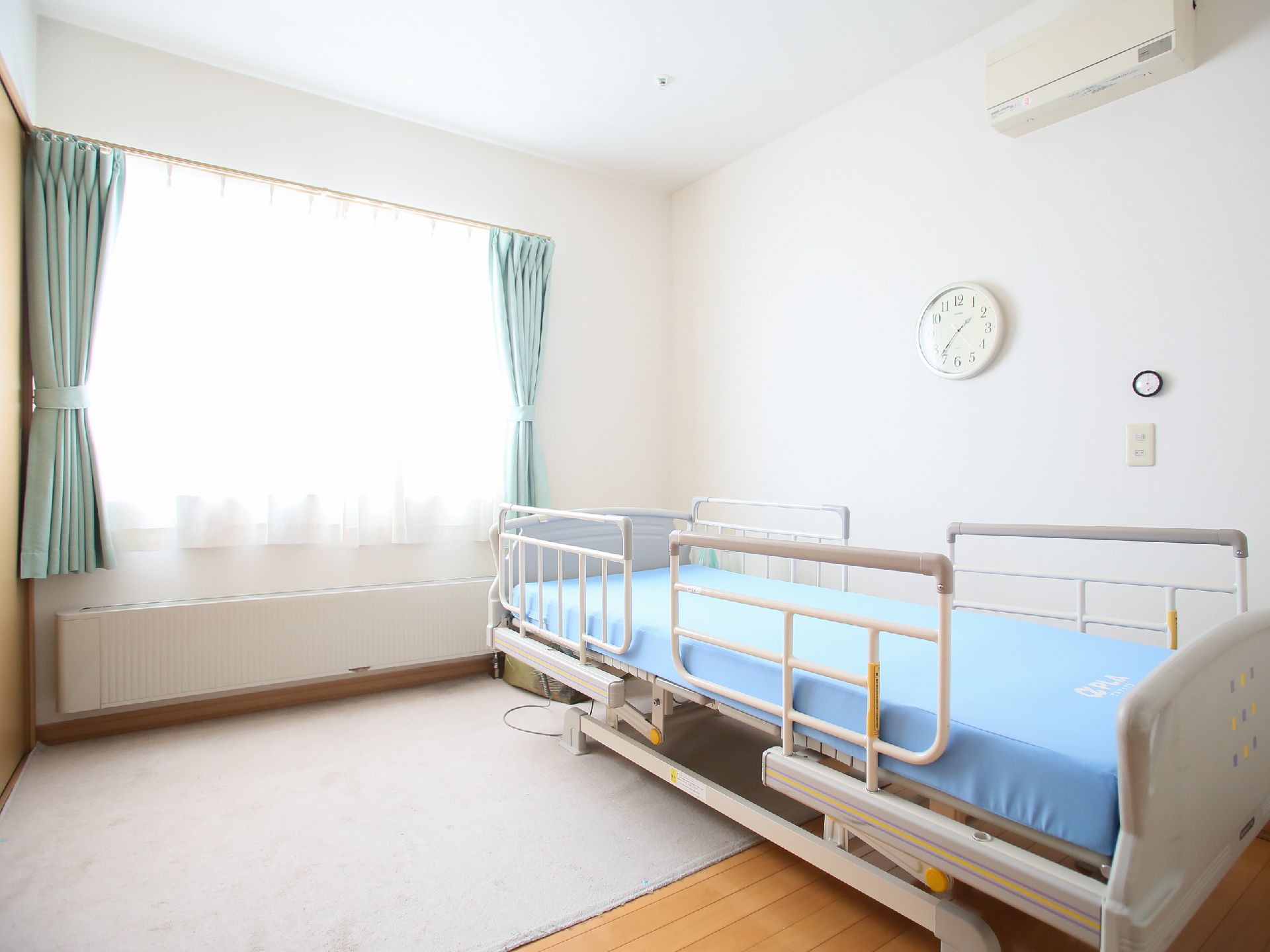 緑のカーテンがかかり、ベッドが置かれている白い内装のシンプルな部屋