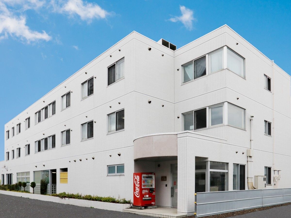 白を基調とした3階建ての建物にはたくさんの窓が付き、玄関の近くには赤い自動販売機が設置されている。