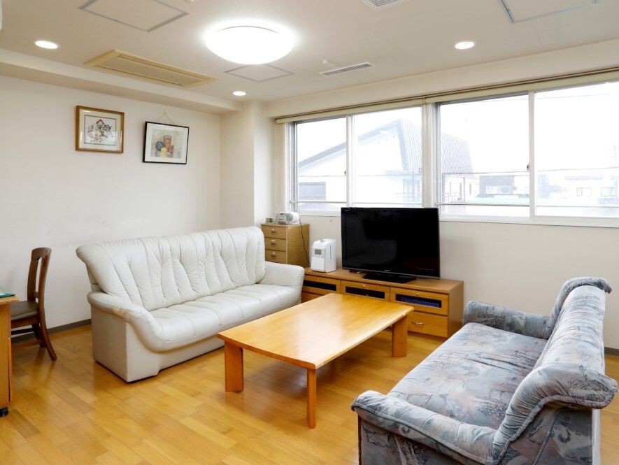 テレビやテーブルを囲んでソファが置かれ、壁には絵画が飾られている。窓の外には住宅地が見える。