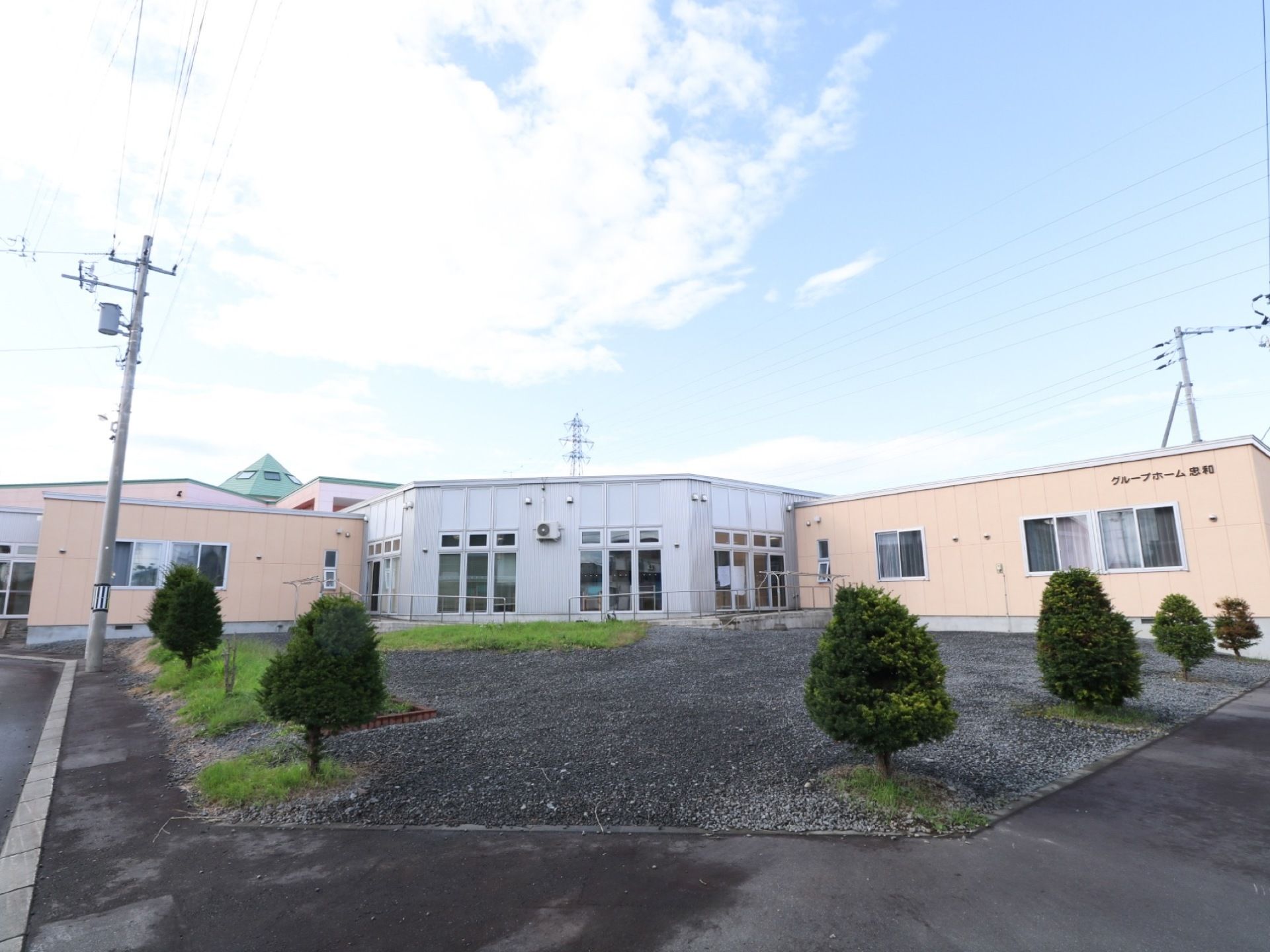 白い建物とベージュ色の建物があり、右側の建物には施設名が書かれている。手前は砂利が敷かれたスペースになっており、低い木が植えてある。