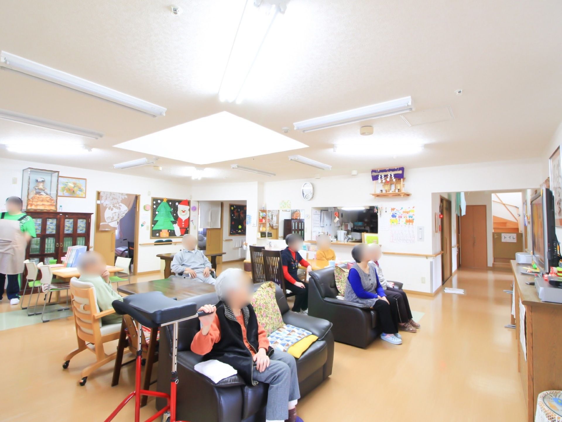 共有スペースに多くの入居者様が集まって座っている。後ろの方に、エプロンをしたスタッフが１名立っている。