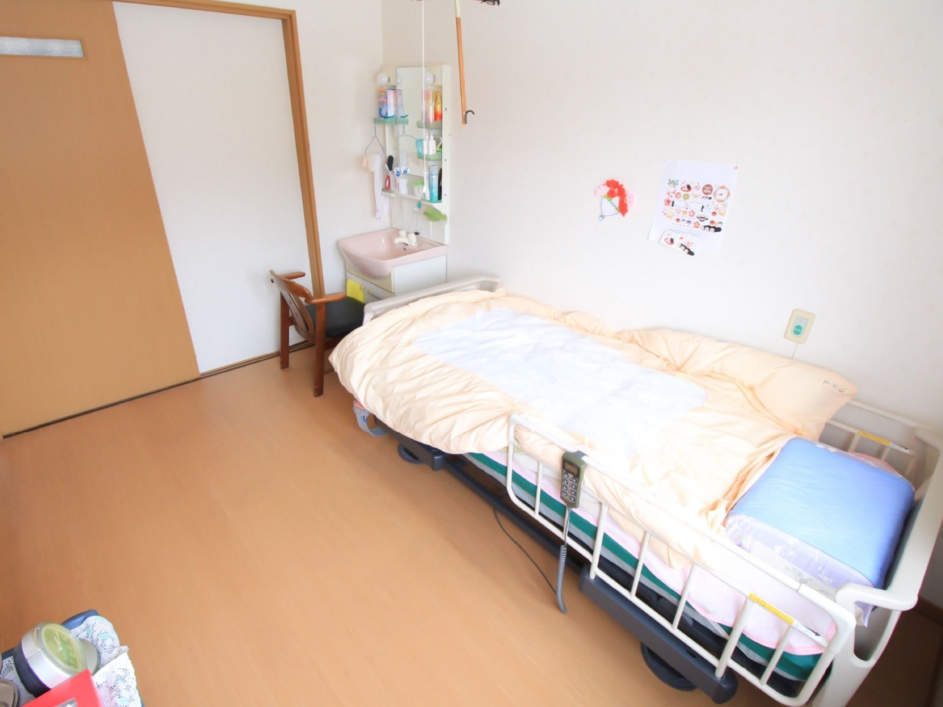 居室の壁際に転落防止柵つきのベッドが設置されている。入口扉のすぐ近くに、ピンク色のシンクの洗面台がある。
