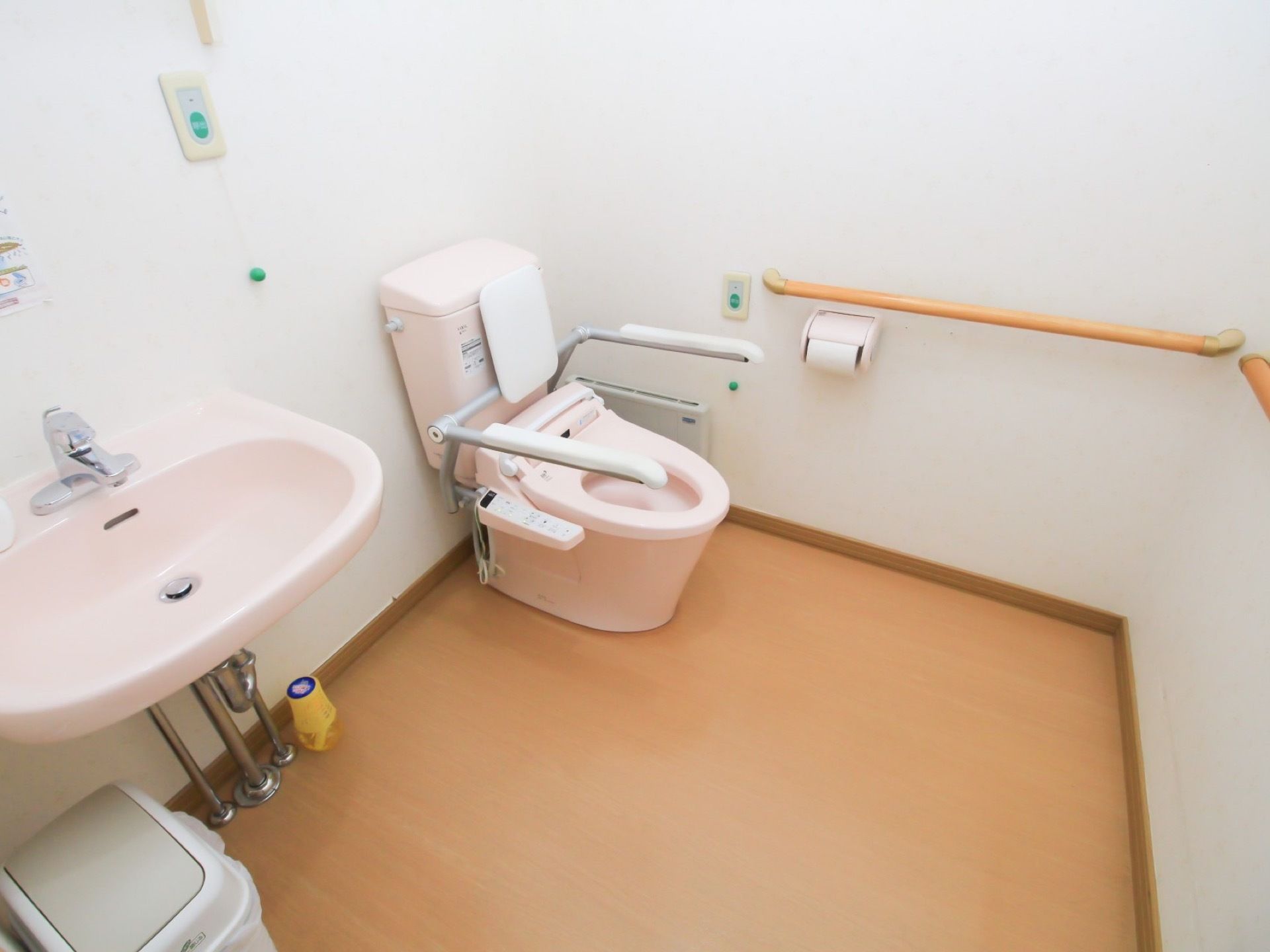 共用トイレには広いスペースがある。壁に横方向の大きな手すりが取り付けられている。便器の横に洗面台が設置されている。