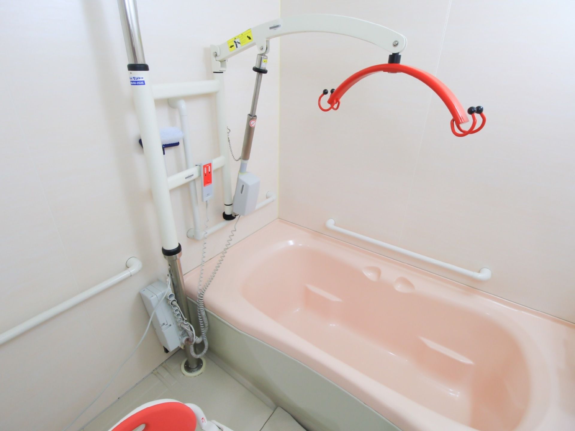 浴室に介護入浴設備が設置されている。赤色のフックでつり上がる形で浴槽に入ることができる。支持棒が浴槽の横に立っている。
