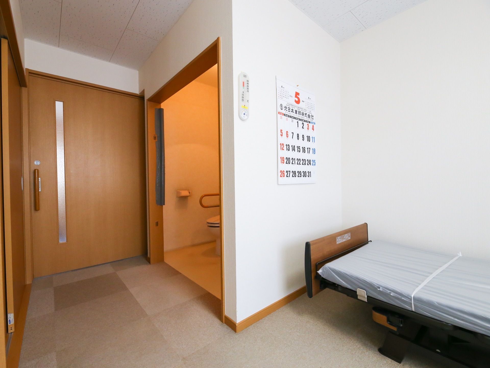 正面に廊下へ出る引き戸の扉があり、その前にトイレが見えている。手前は居住スペースになっており、介護用ベッドが置かれている。