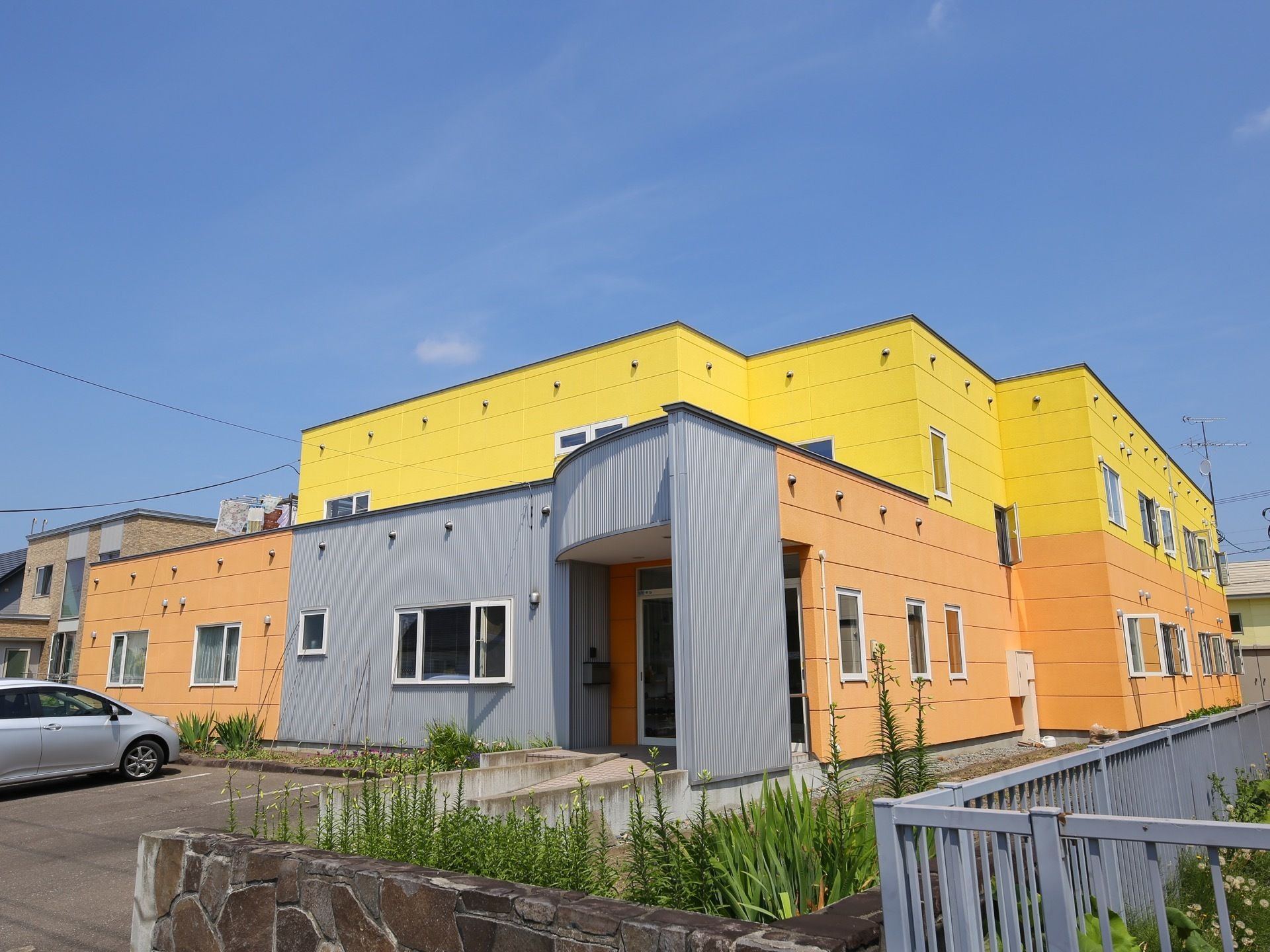 オレンジやグレー、黄色に塗り分けられた外観の建物で、２階建てになっている。玄関前に駐車場が広がっている。