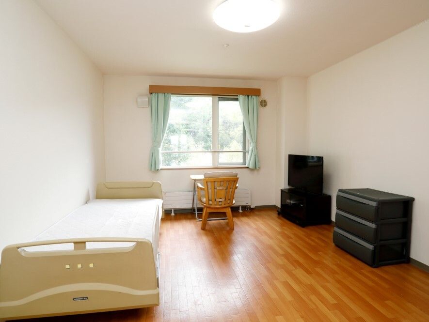 窓際にはテーブルセットが置かれ、寝具付きのベッドやタンスなどもあるフローリングの部屋。大きな窓があり明るい。
