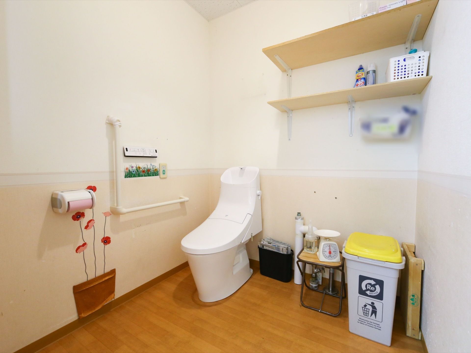 トイレの壁には、２枚の棚が取りつけてあり掃除用具などを整頓して置いてある。壁には、花のシールが貼ってあり、明るい印象である。