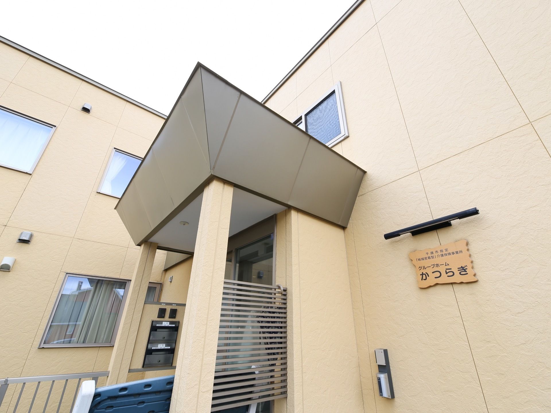 ベージュの外壁から、玄関前にグレーの屋根が張り出している。屋根の下に、２台のインターフォンがついている。