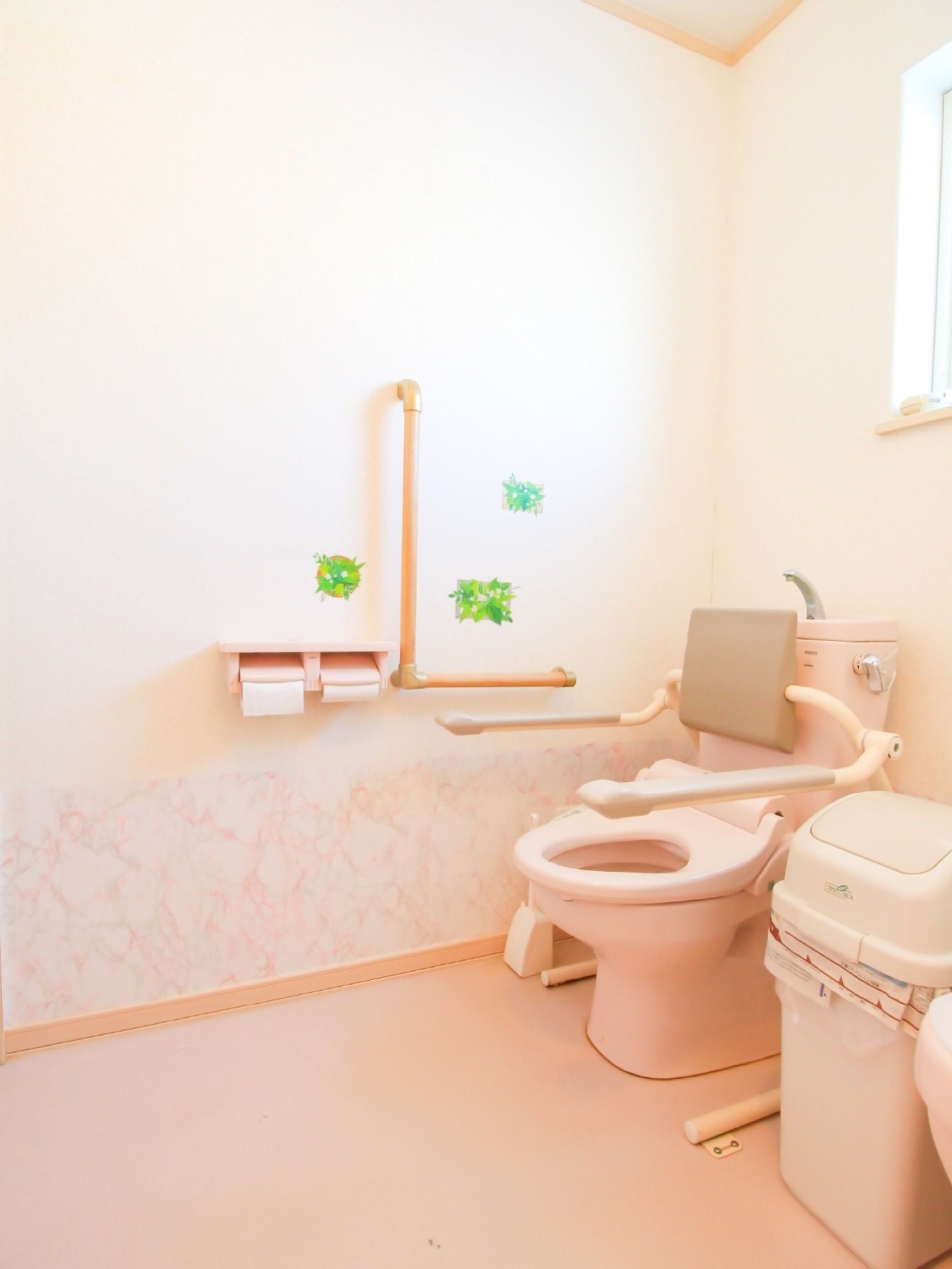 広いスペースを確保されたトイレである。トイレ用手すりが付けられており、立ち座りに便利である。緑の壁紙が貼られている。