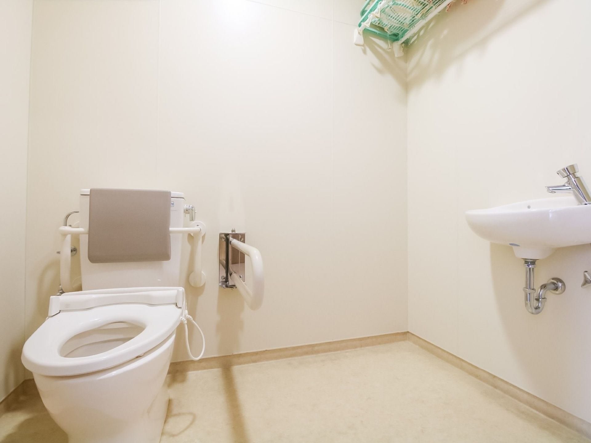 共用トイレの個室内の便器には、背もたれと手すりが設置されている。シンプルな個室で、小さな手洗い場と突っ張り棒の戸棚がある。