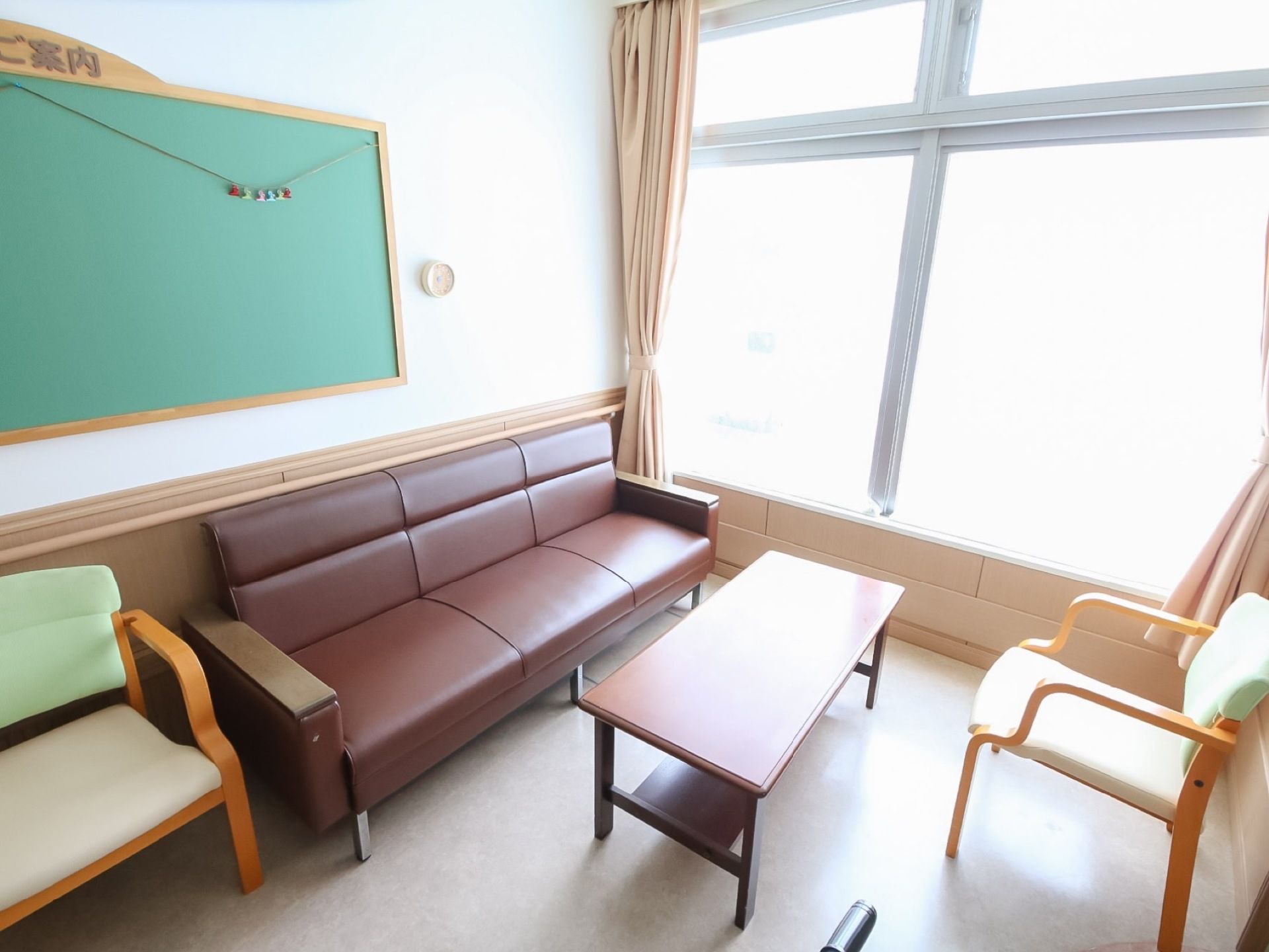 窓際の明るい場所に、シンプルなテーブルとソファー、椅子が並んでいる。壁には「ご案内」と書かれた掲示板がある。