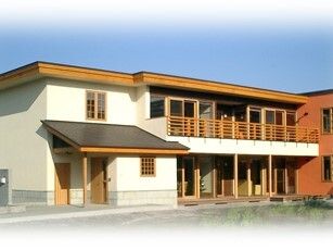 家庭的な雰囲気が漂う2階建ての建物。白やブラウンを使っていて、玄関には大きな屋根があり、2階部分にはテラスが設置されている。