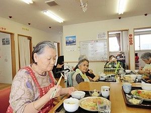 赤いエプロンを付けた女性入居者が手前に座っている。ダイニングテーブルの上にはトレーに並べられた食事がある。