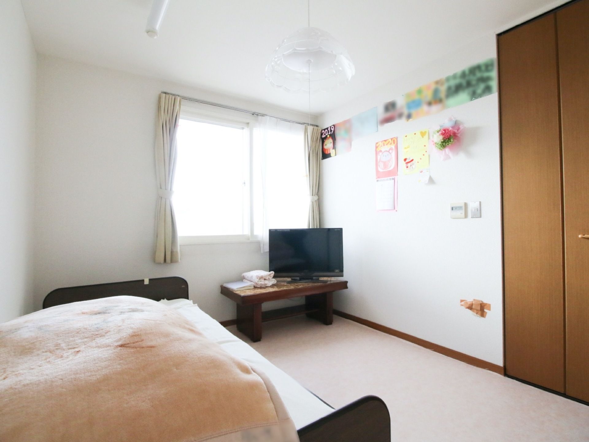 窓際に茶色のミニテーブルがあり、その上にテレビが設置されている。左側にはベッドが置いてあり、毛布が掛けてある。