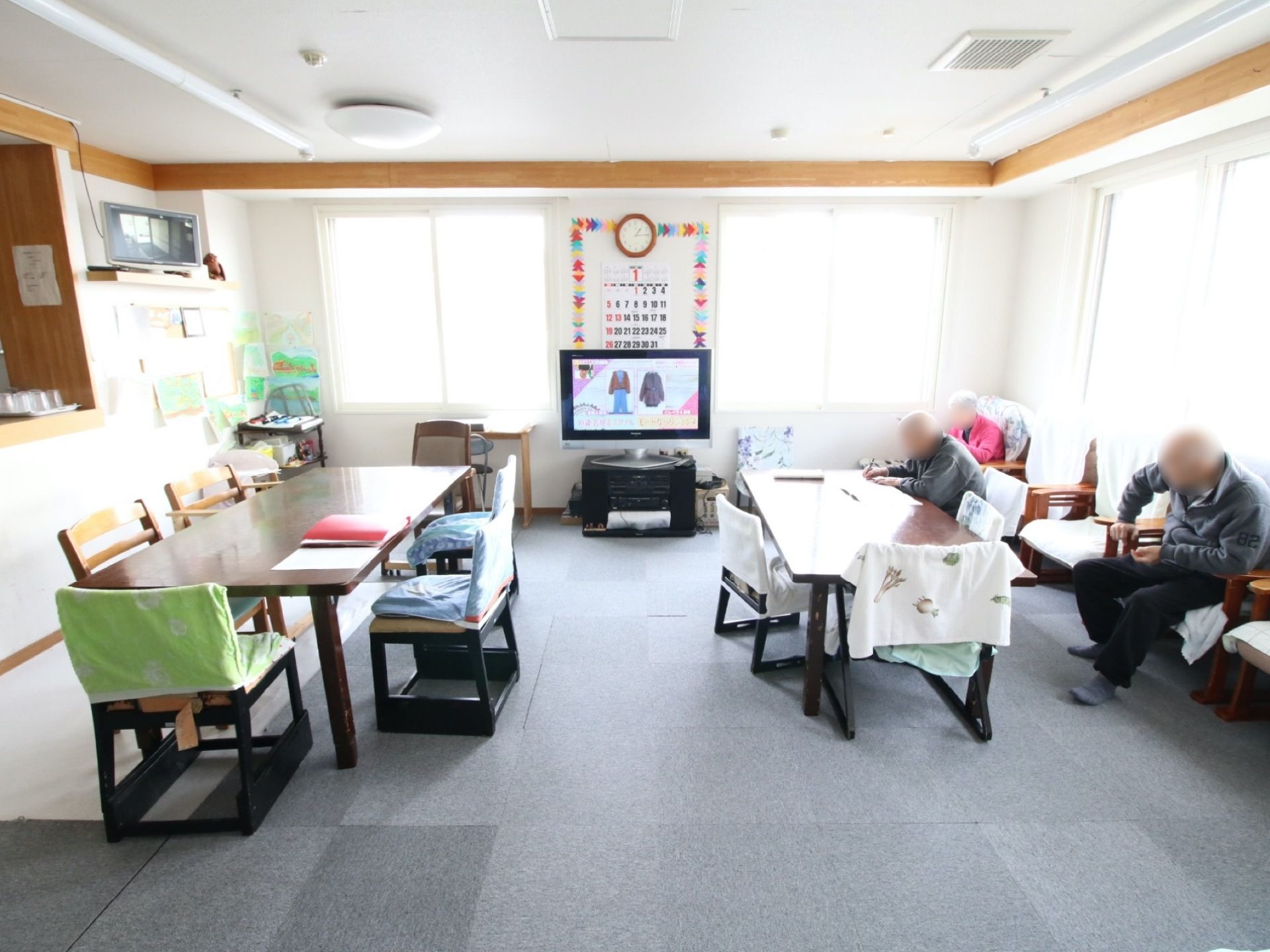 大きなテレビが壁際に置いてあり、その後方にカレンダーが掛けてある。数名の入居者様が、テレビを見ながらくつろいでいる。