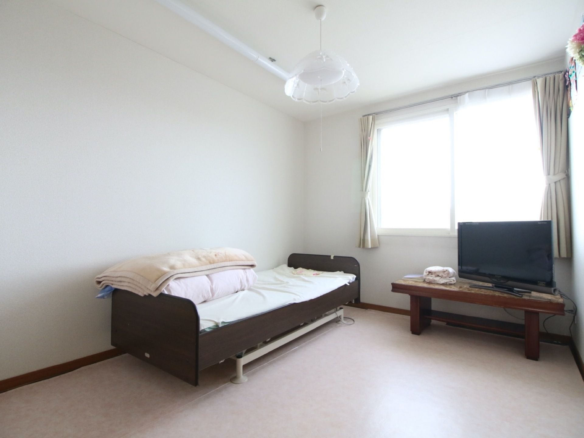 居室内に置いてあるベッドや机は、茶色系の落ち着いた色でまとめられている。天井から、白い照明器具が吊り下げてある。