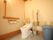 ベージュ色の背当てと、可動式の手すりが付いたトイレである。左側の壁にもＬ字型の手すりがあり、その下に操作パネルがある。