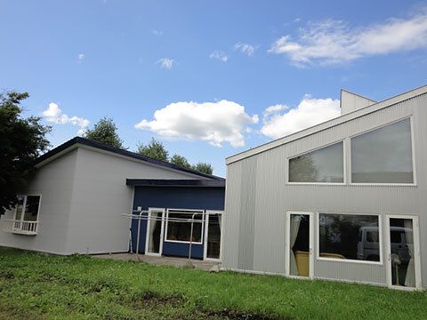 青空の下に建っているブルーとグレーの建物。敷地内には洗濯物干し場もあり、畑のような場所もある。