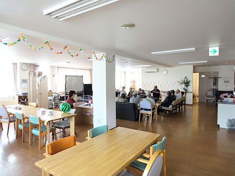 手前には木製のダイニングテーブルがあり、奥のソファスペースに入居者が集まってレクリエーションをしている。