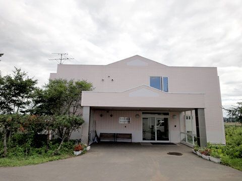 白っぽい外壁の建物は３階建てで、窓が少なく、玄関には車寄せが付き、ベンチが置かれている。