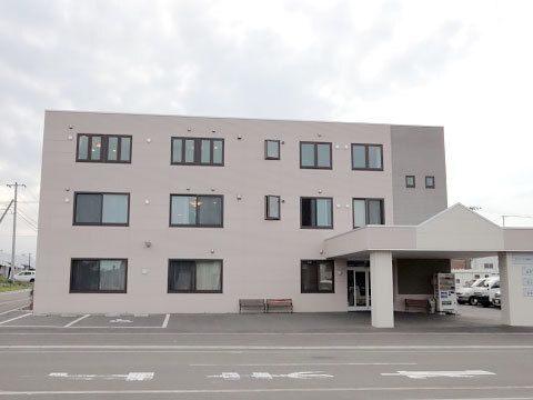 周りには建物がなく、駐車場が付いたグレーの3階建ての建物が建っている。玄関横にはベンチがあり、車寄せも付いている。