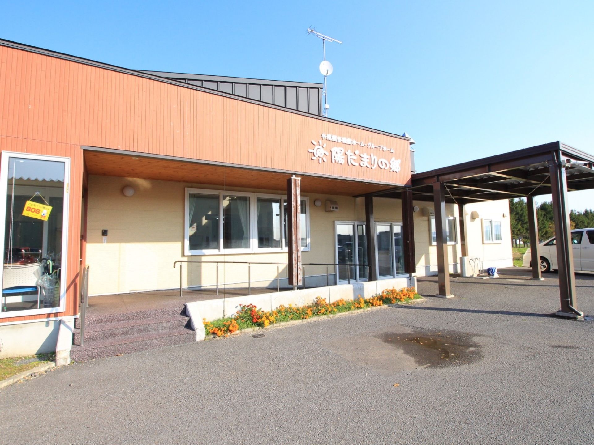 施設は平屋建ての建物で、玄関へのアプローチはスロープと階段である。建物の前に花壇があり、花が咲いている。