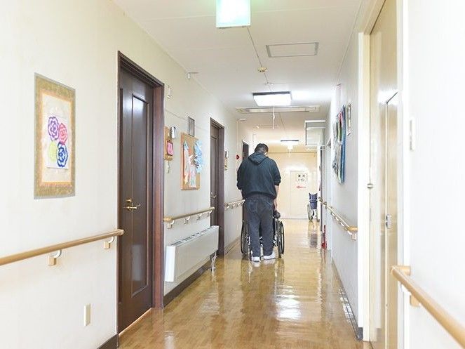 廊下を車いすの入居者を押しながら歩く男性スタッフの後ろ姿。