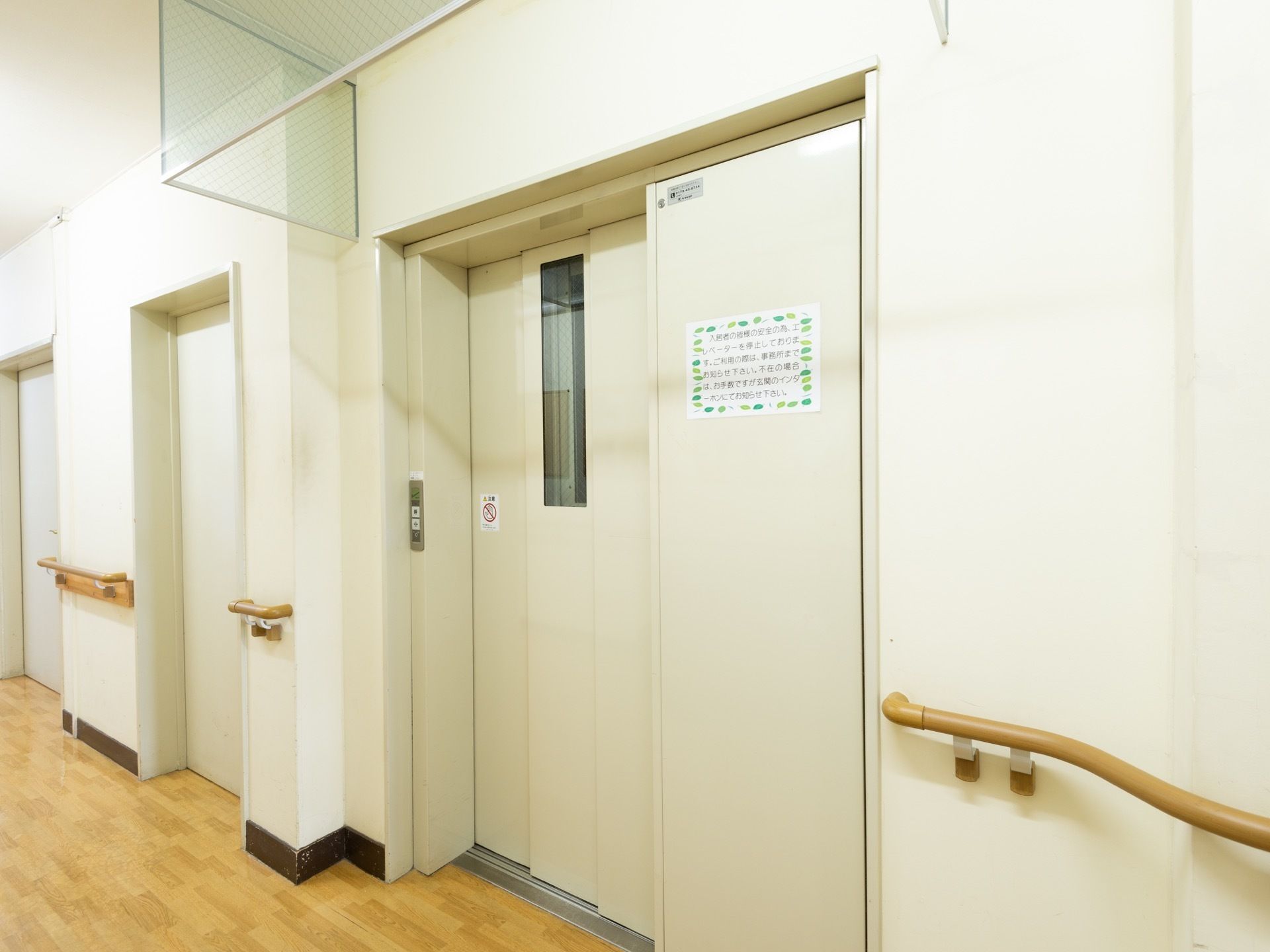 白い壁に茶色の床で、壁には手すりがあり、安全に移動することができる。右側には片開きの扉のエレベーターが設置されている。