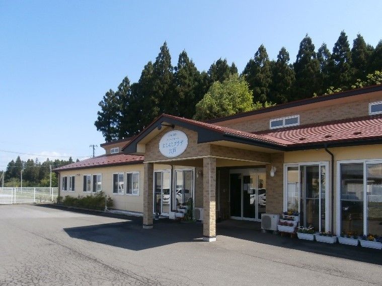平屋建ての建物は明るい色でまとめられ、玄関前にはたくさんのプランターが置かれ花が咲いている。