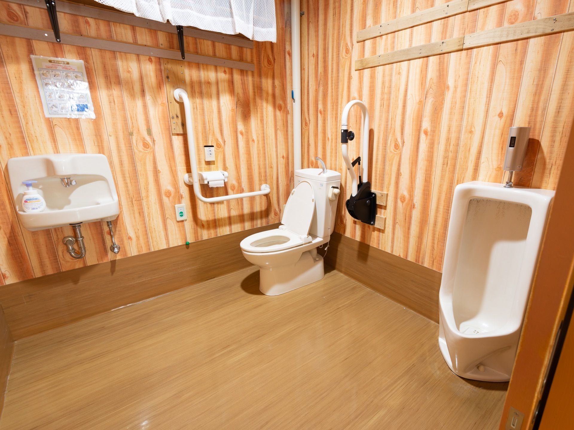 木板の壁に囲まれた共同トイレがある。男性用と共用の便器が設置されている。洗面台にはハンドソープのボトルが置かれている。