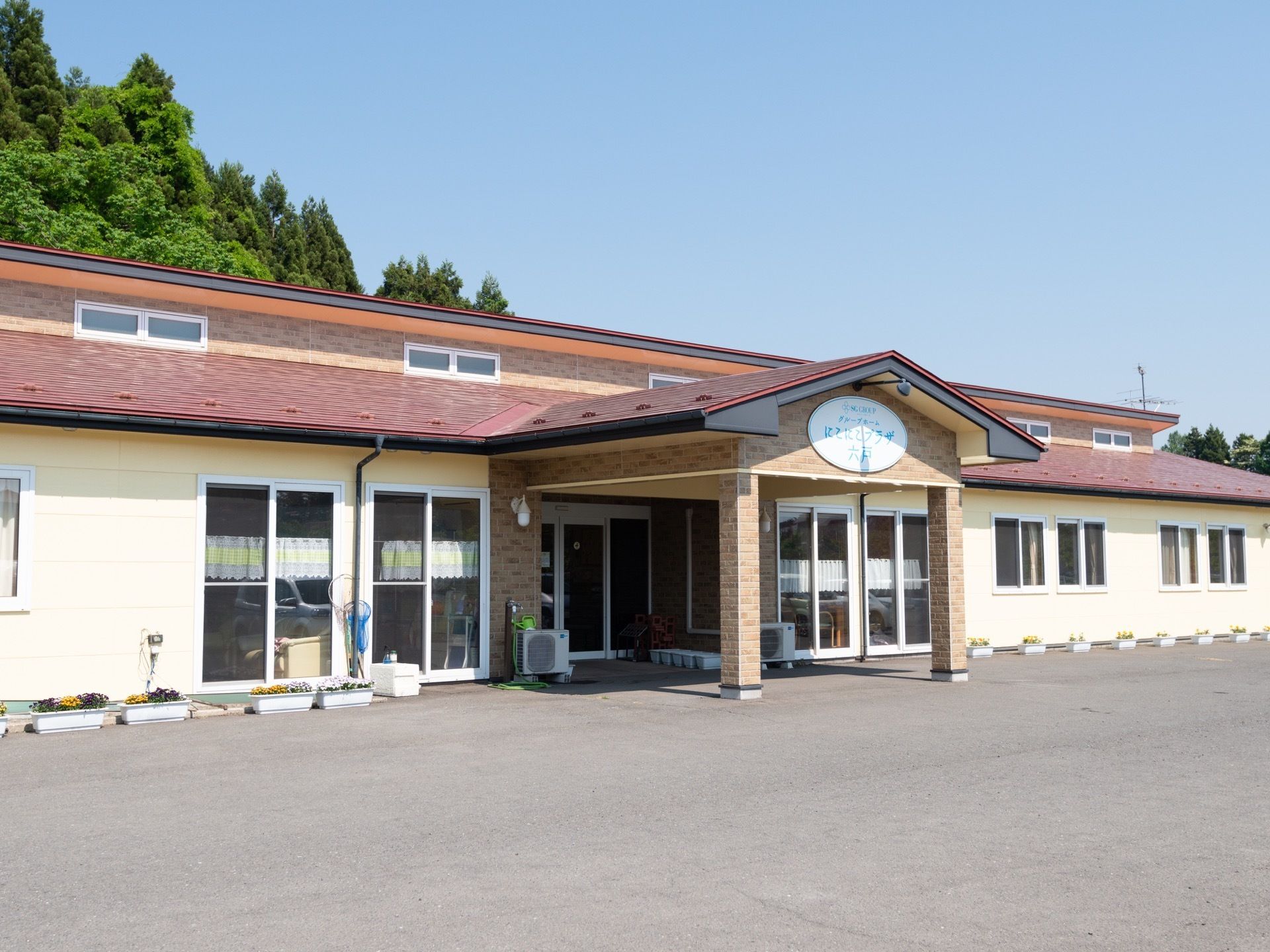 建物の外壁は、あたたかみのある色合いのレンガや塗装が使われている。施設の裏手には、森林が見える。建物の前面には、広いスペースがある。