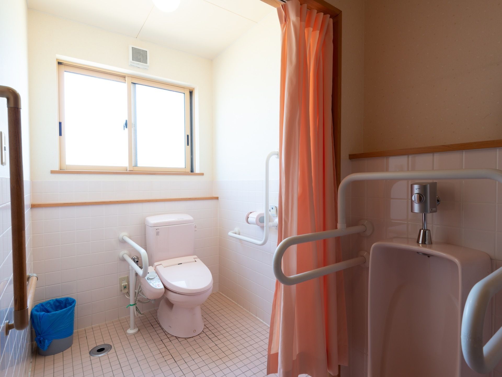 開放的なトイレである。小便器も設置されている。手すりが備え付けられており、壁にはペーパーホルダーが備わっている。
