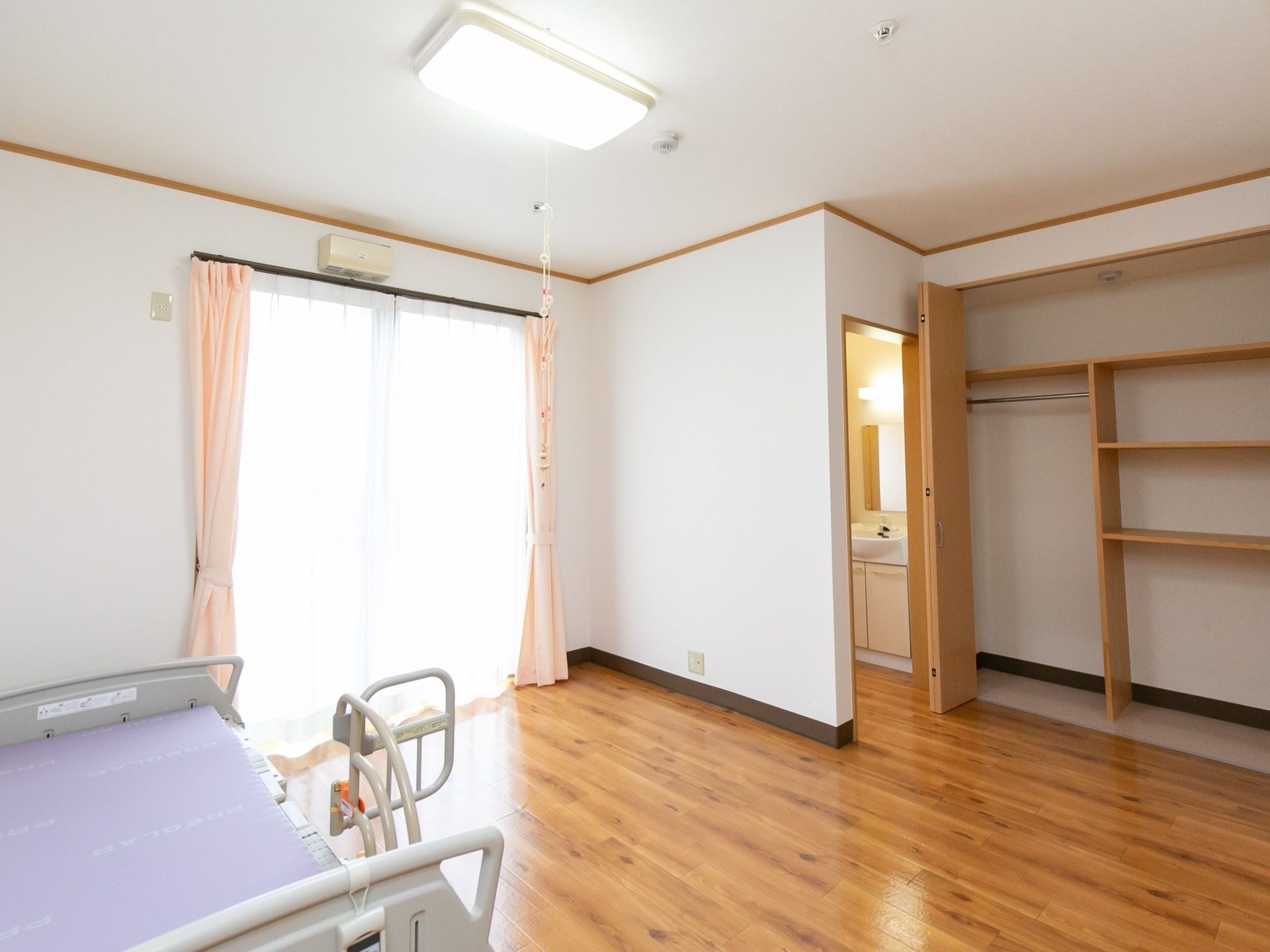 フローリング張りの床に、白い壁の居室である。左手前に介護用ベッドが置かれている。右奥には折れ戸扉のついた収納スペースがある。