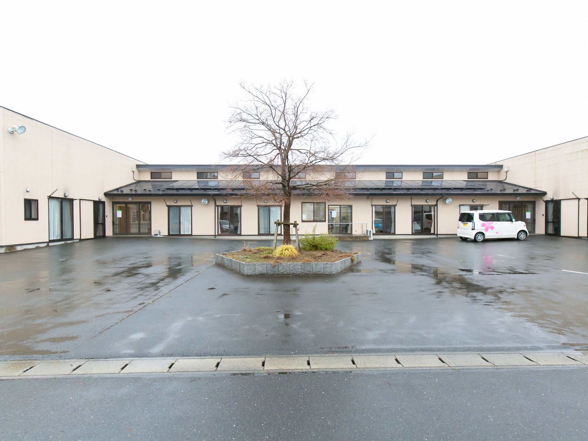 施設前の通りから玄関周りを見た画像である。正面に平屋造りの施設が建っている。施設の正面は広い駐車スペースになっている。