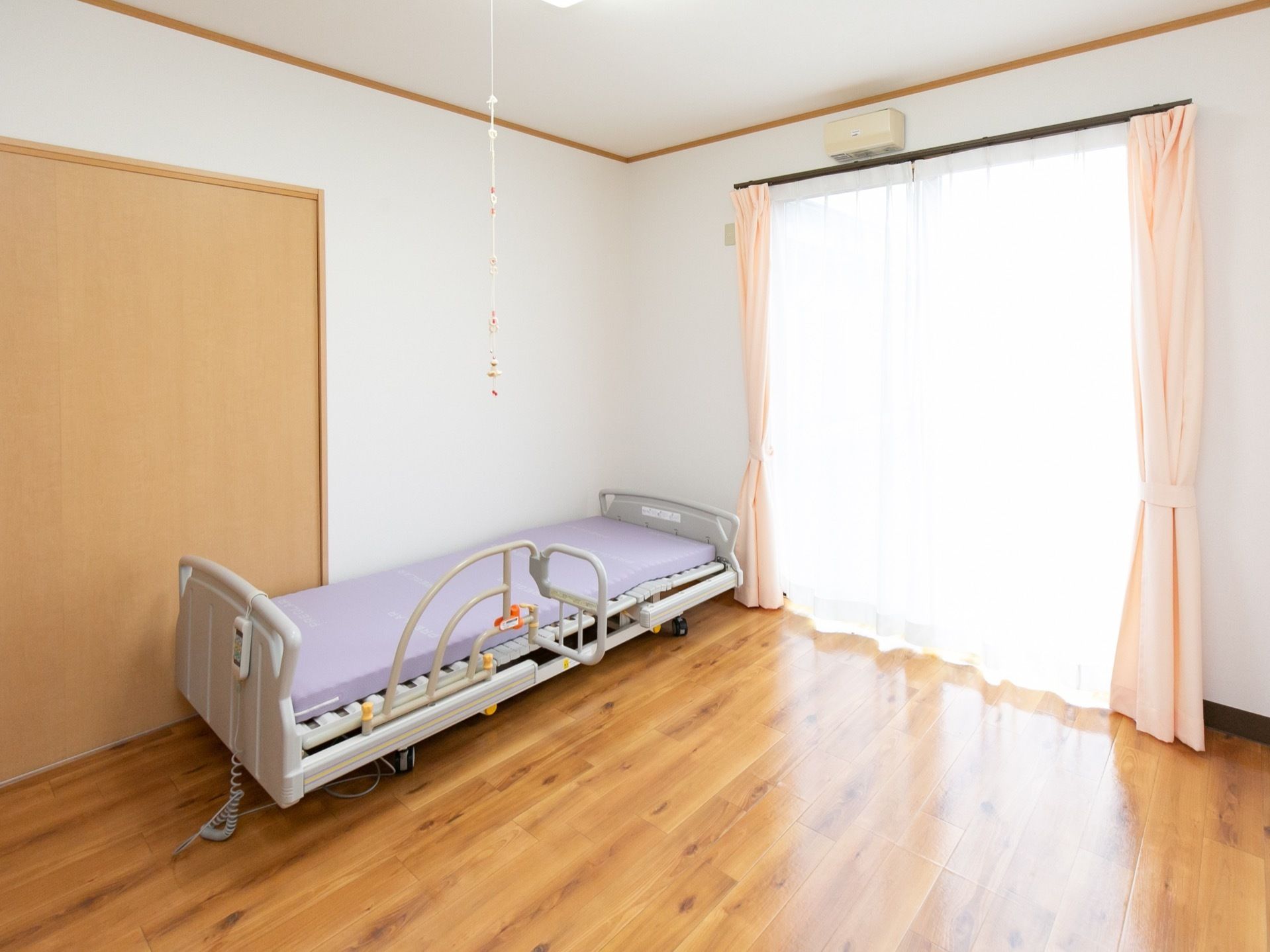 床はフローリングで、右奥に外へ出られる大きな窓がある。薄いオレンジのカーテンが束ねられている。壁際に介護用ベッドがある。