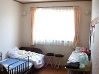 大きな窓がある小さ目のワンルームタイプの部屋。介護用ベッド、日用品が置いてある