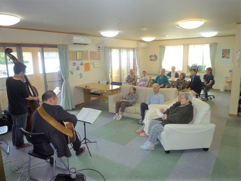 ギターなどの還元楽器を弾いている男性二人を囲むようにしてソファが置かれ、入居者が熱心に聞いている様子。