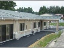 グリーンの屋根が付いた平屋建ての建物。外壁はベージュで周辺には民家もある。