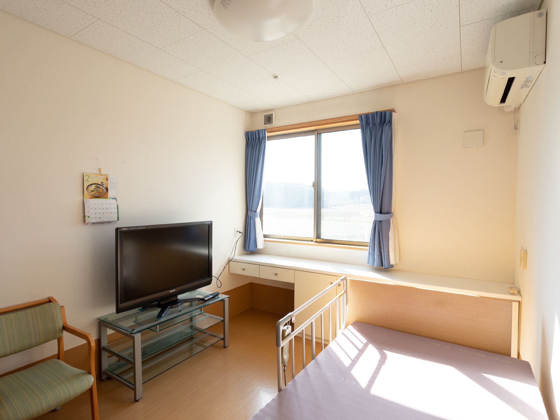 緑の多い屋外を笑顔で散歩しているエプロンを付けた若い女性スタッフと車いすの高齢女性。